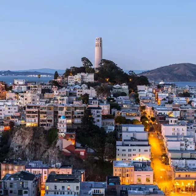 San Franciscos Coit Tower in der Abenddämmerung, mit beleuchteten Straßen davor und der Bucht von San Francisco dahinter.